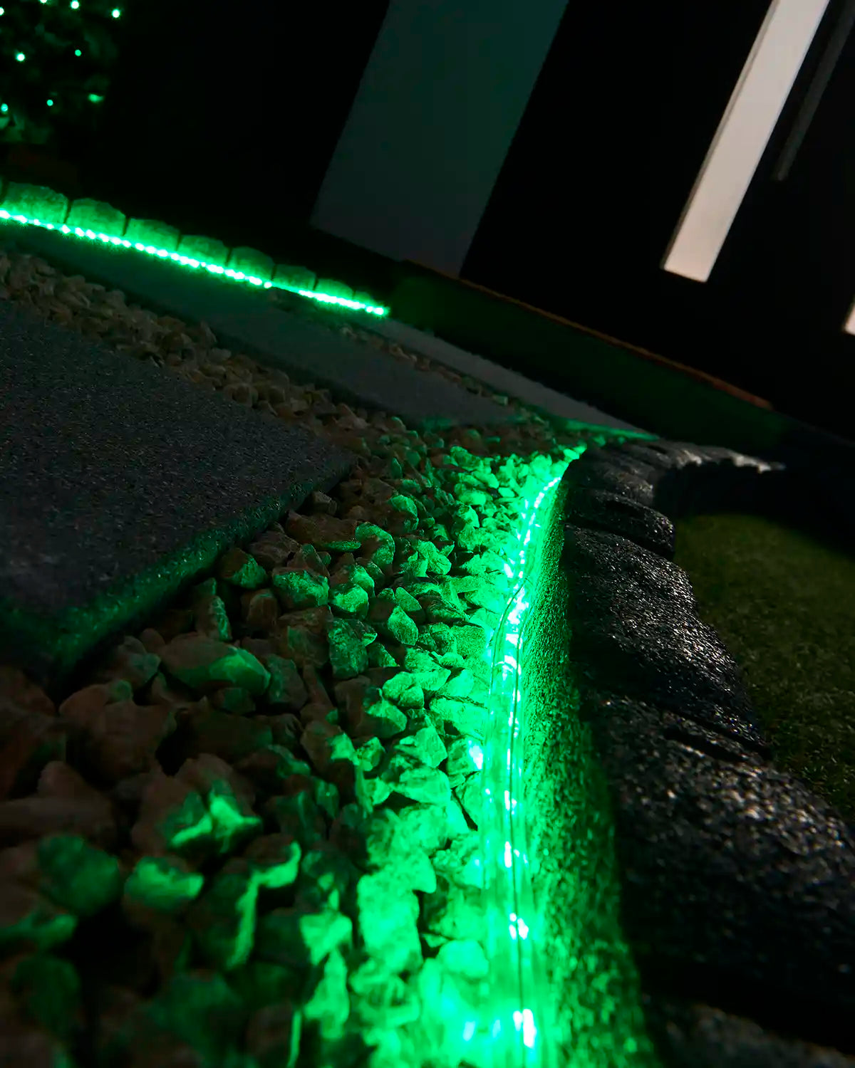 Green rope light illuminating a stone border beside outdoor paving and gravel.
