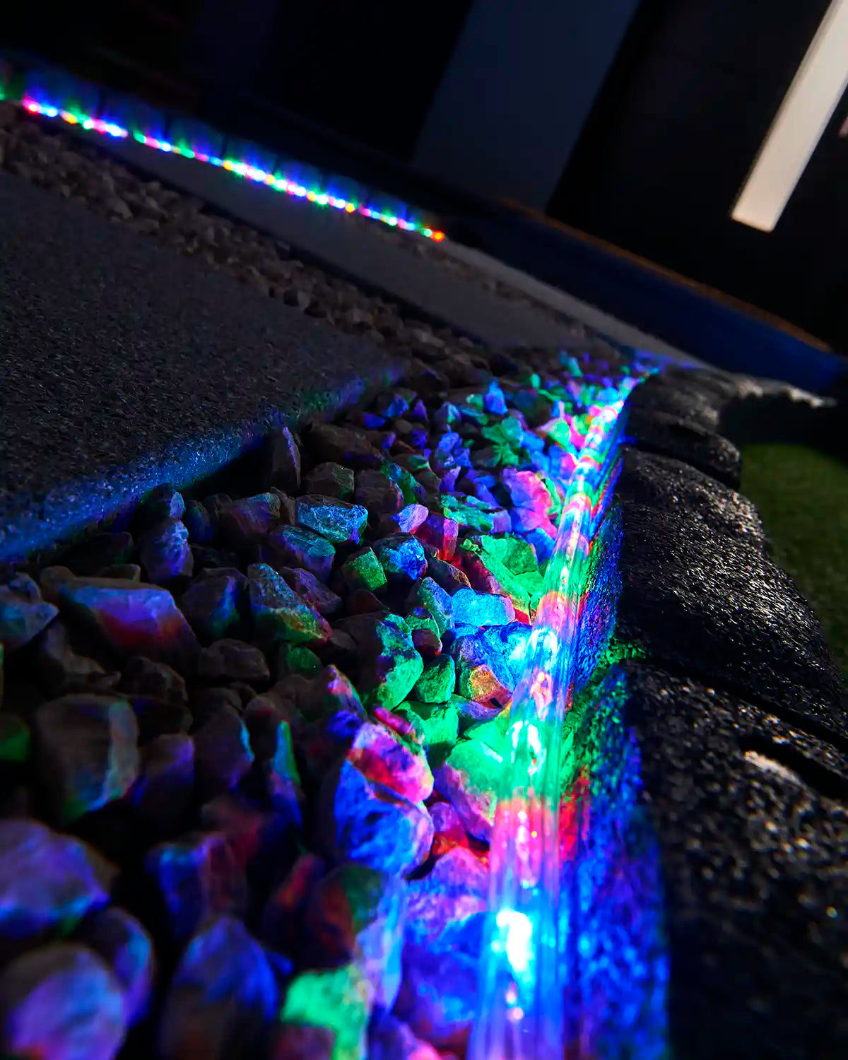Multi colour rope light illuminating a stone border beside outdoor paving and gravel.