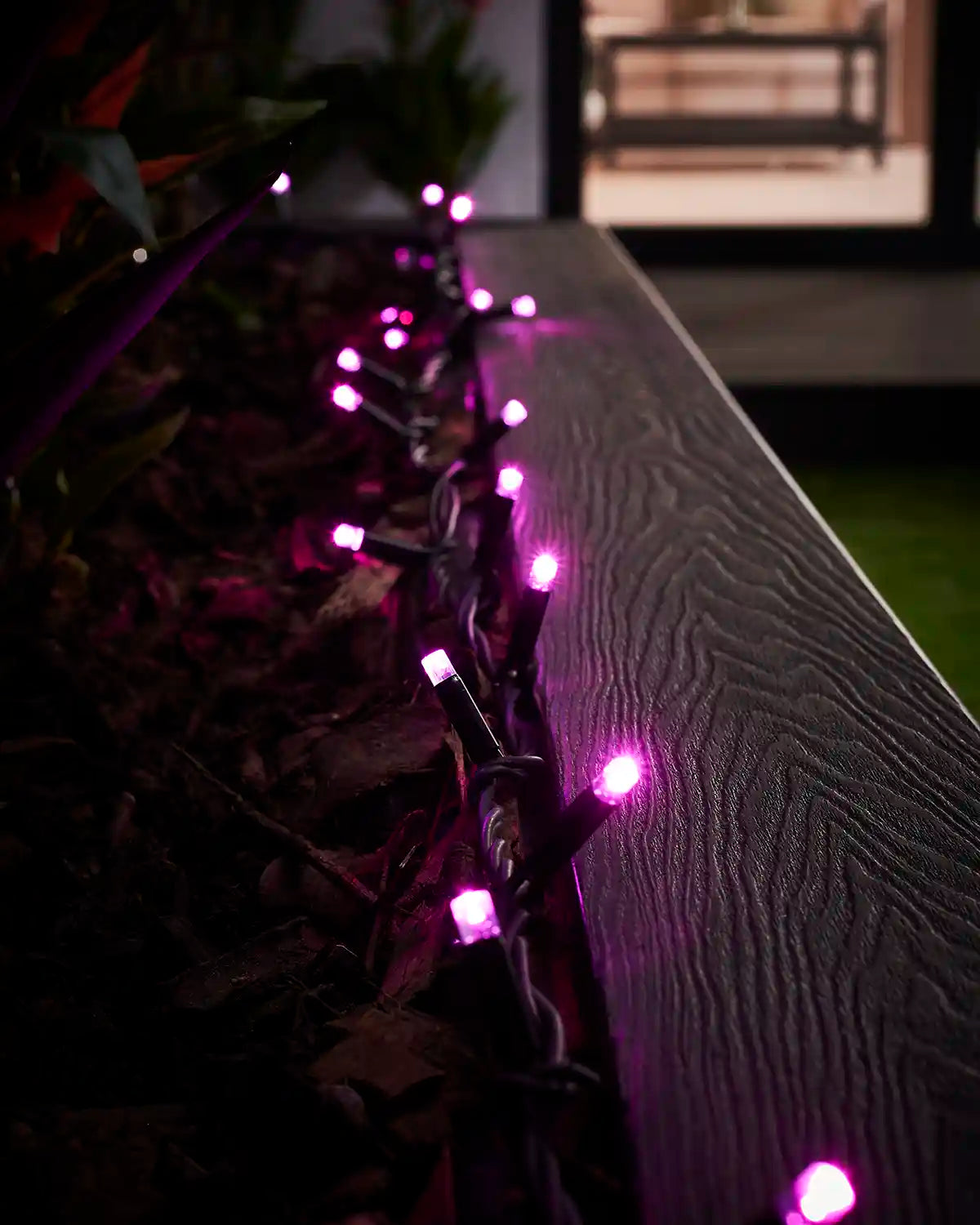 Pink LED string lights on black cable running along an outdoor planter rail.