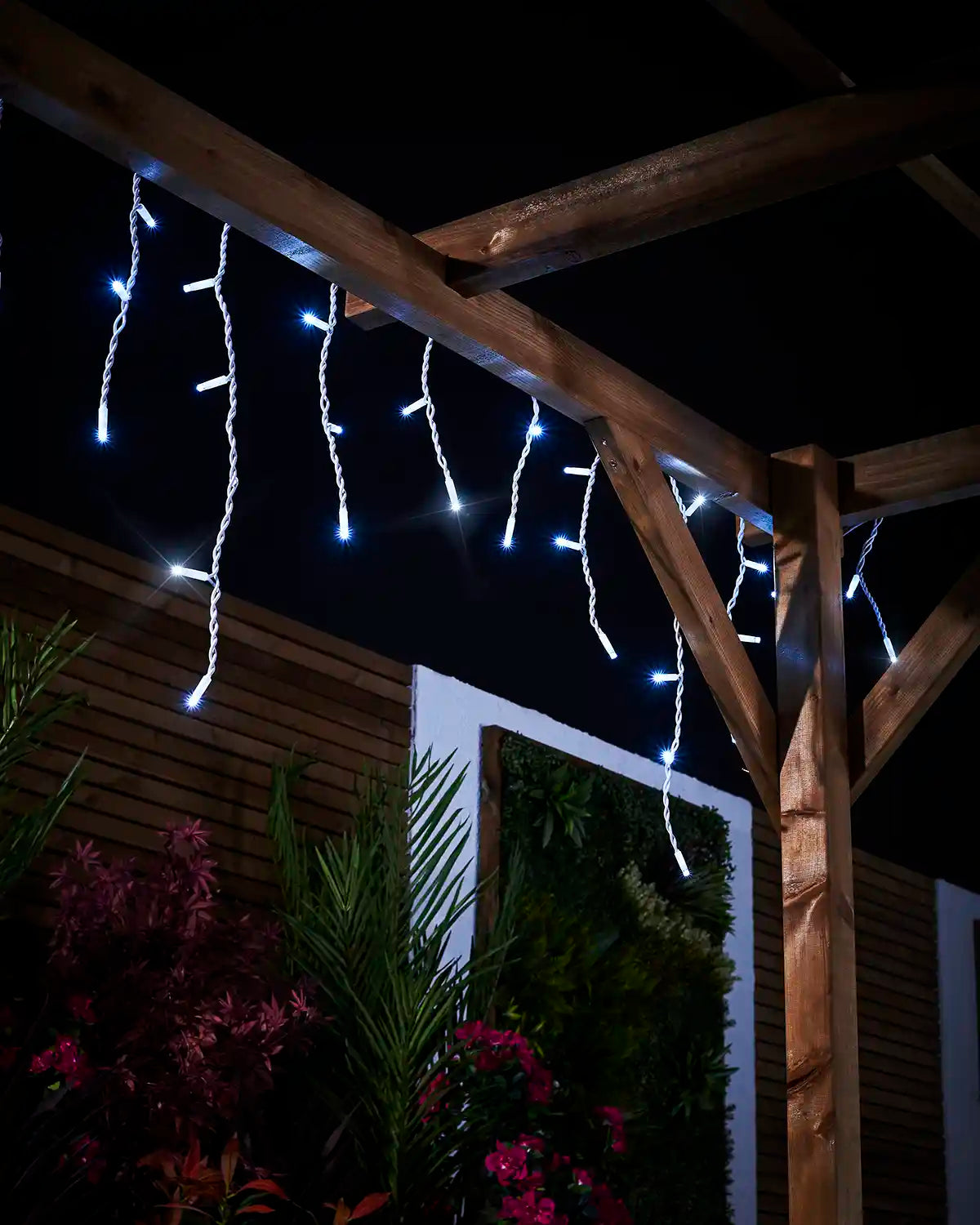 White icicle lights hanging from a wooden pergola above garden plants.