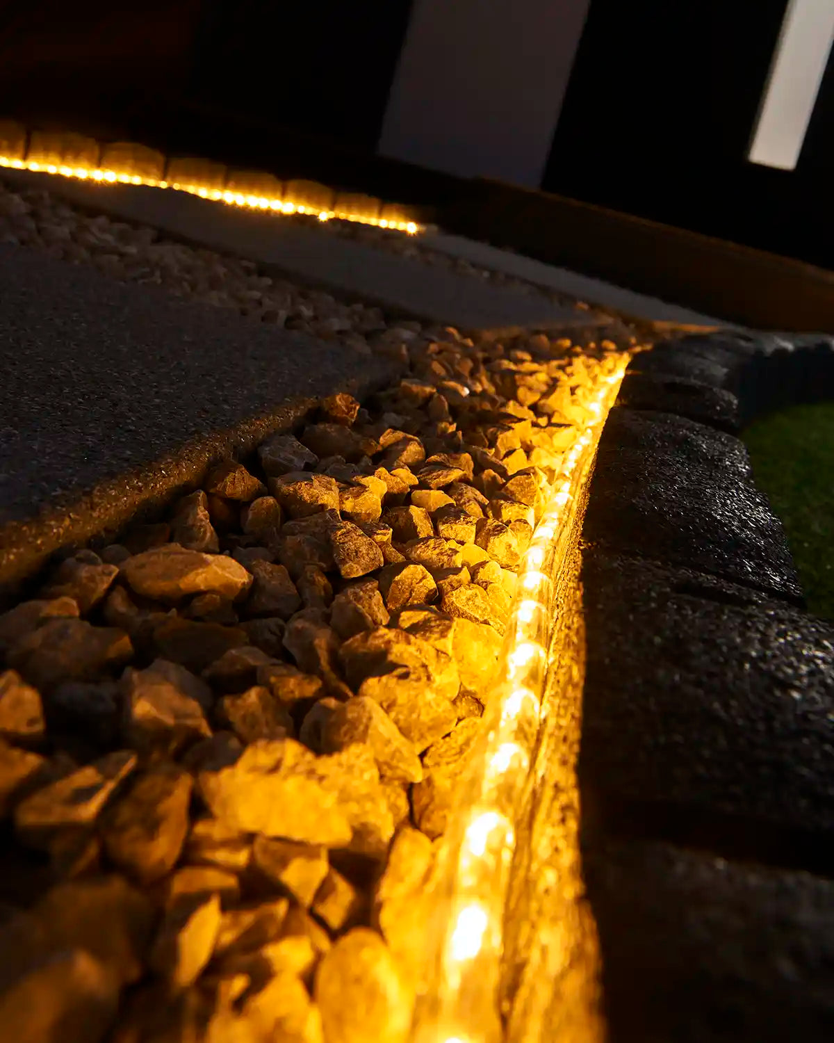 Warm white rope light illuminating a stone border beside outdoor paving and gravel.