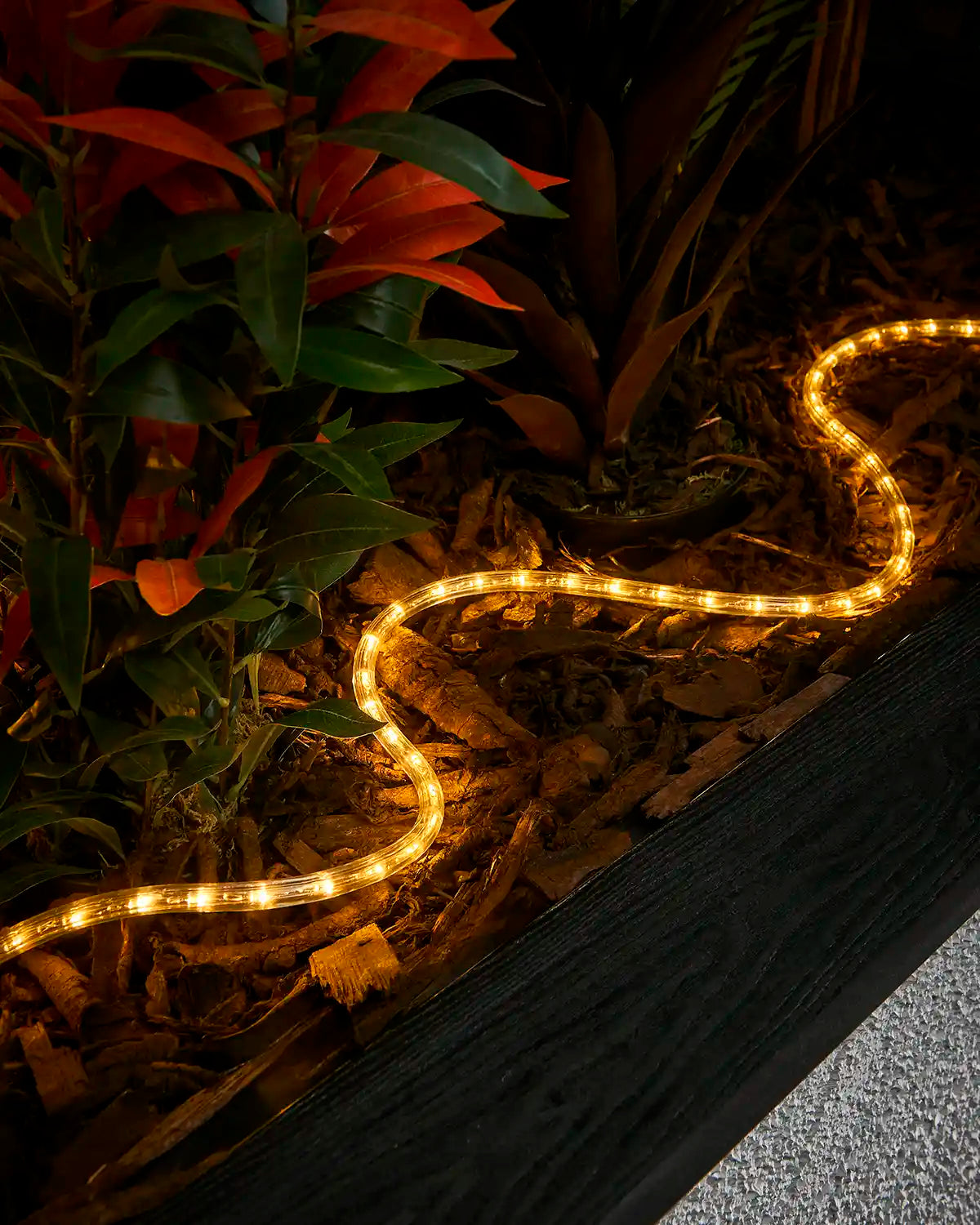 Warm white rope light laid through bark around plants beside a raised garden border.