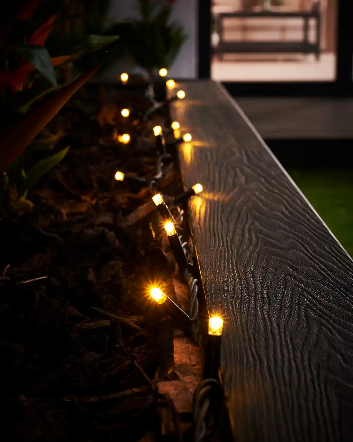 Warm white LED string lights on black cable running along an outdoor planter edge.