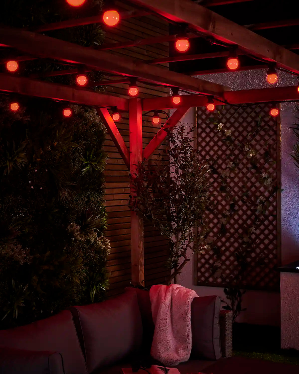 Close-up of red E27 festoon bulbs glowing beneath a wooden pergola with trellis and outdoor seating below.