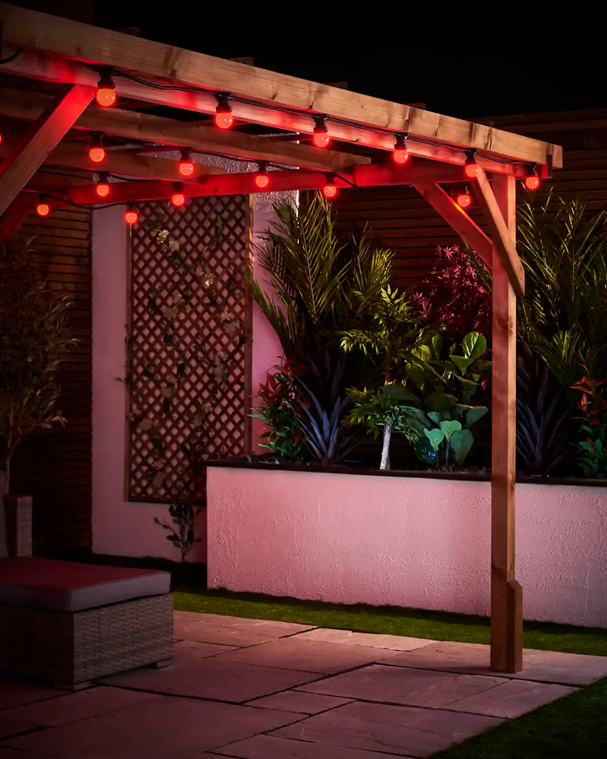 Red E27 festoon bulbs hanging from a wooden pergola above a paved patio with a raised garden bed behind.
