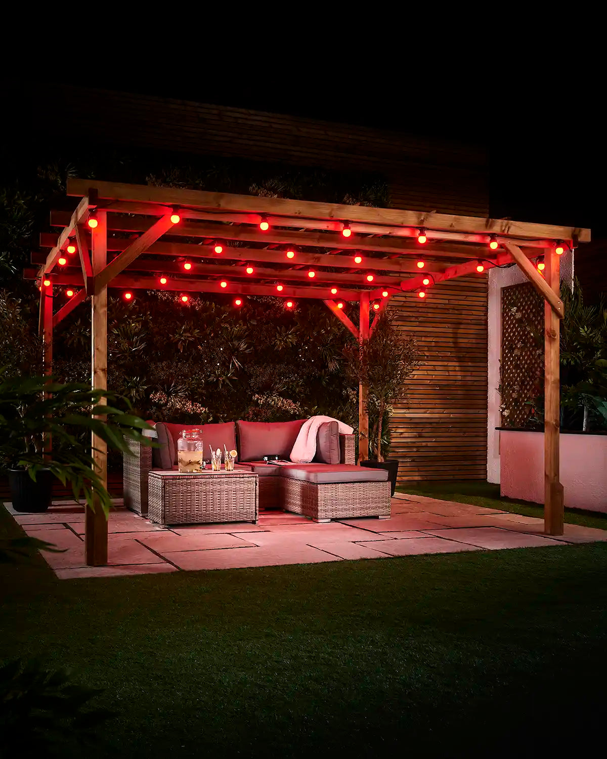 Red E27 festoon bulbs displayed across an outdoor wooden pergola at night with patio seating and garden plants.