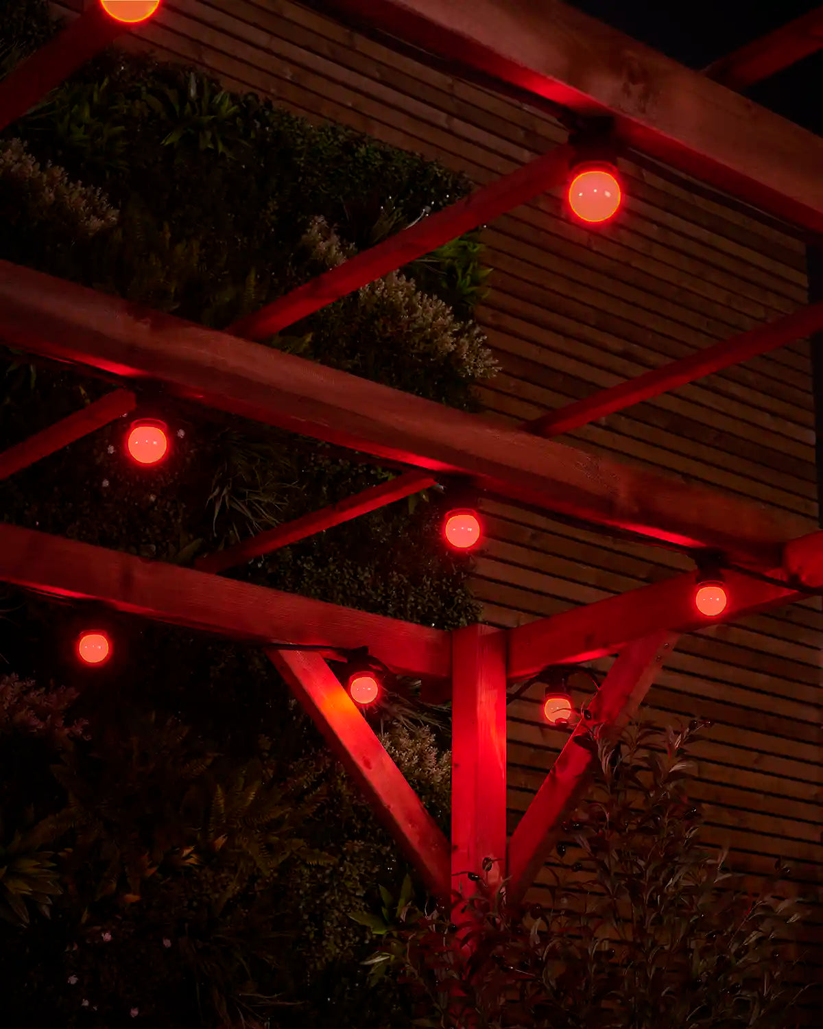 Red E27 festoon bulbs glowing under wooden pergola beams with garden foliage in the background.
