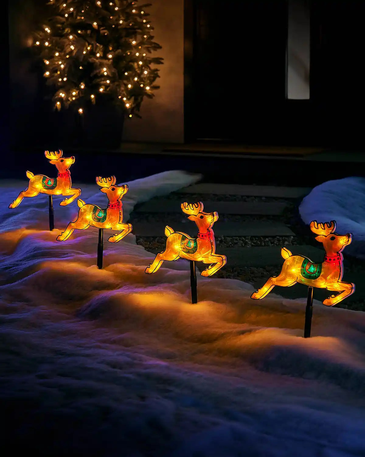 Set of four illuminated reindeer stake lights displayed in snowy outdoor ground near a doorway