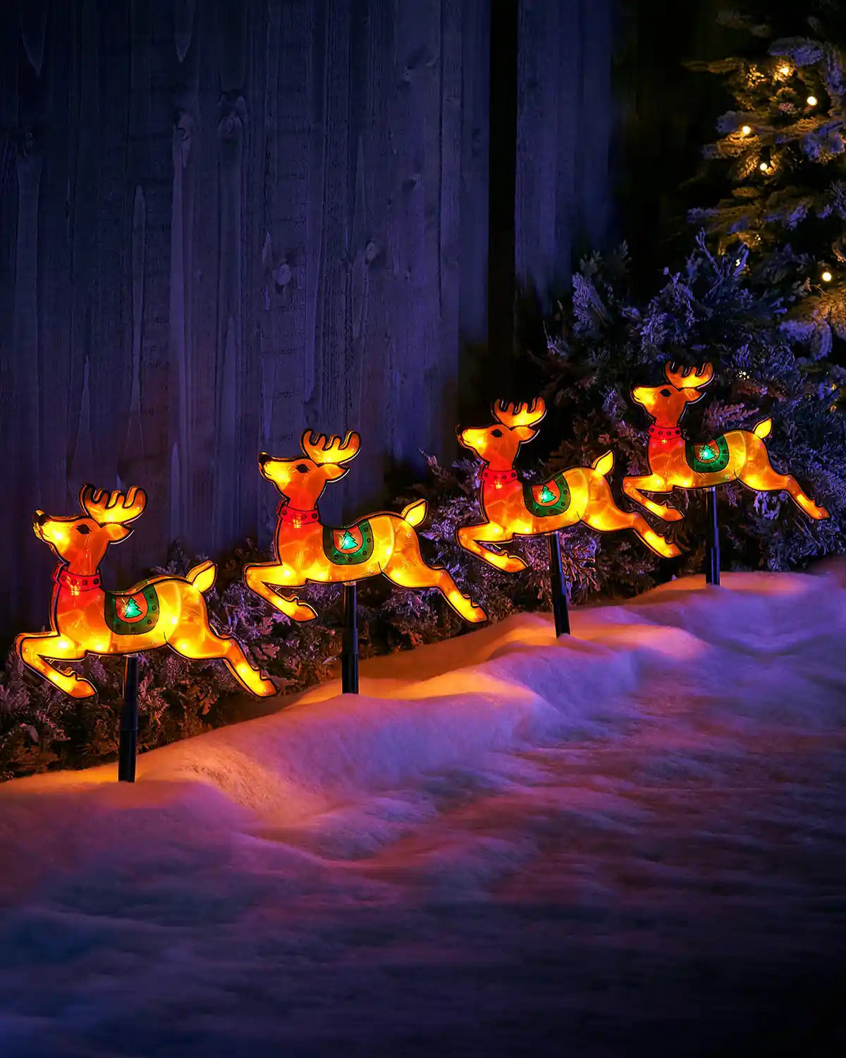 Four glowing reindeer pathway stake lights displayed in snow beside outdoor greenery