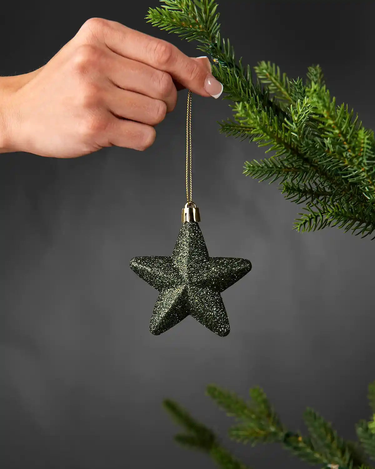 Hand holding a sage green 7cm glitter star decoration beside a Christmas tree.