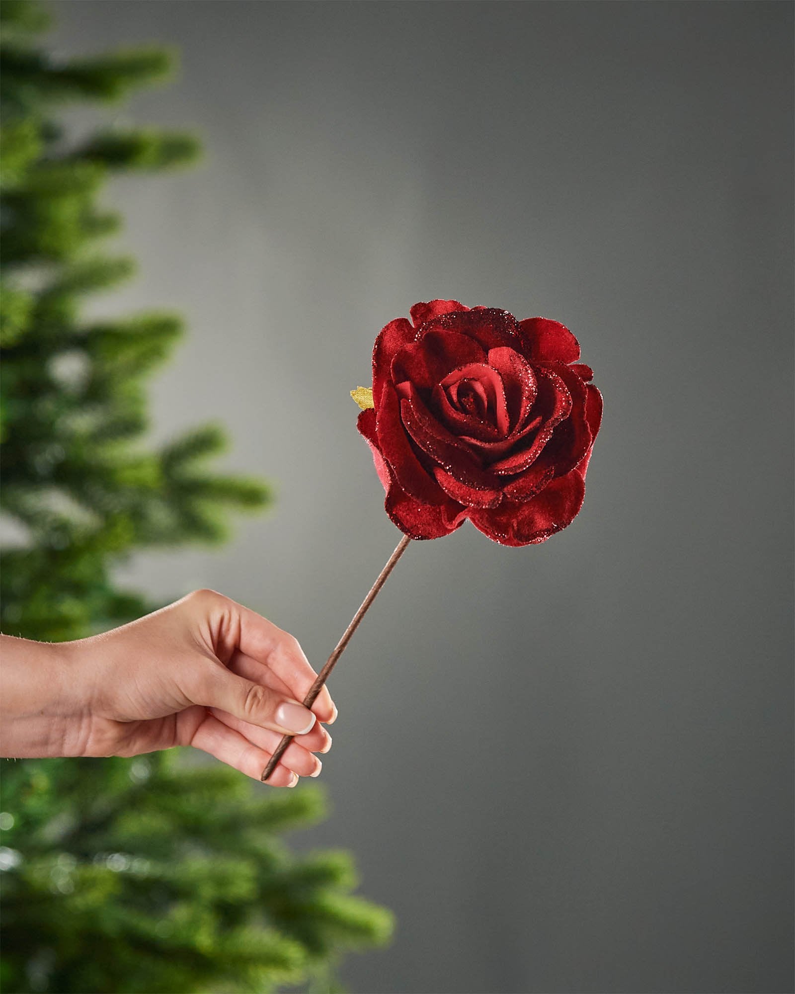 Red artificial glitter rose with layered petals and a brown stem, shown held in front of a Christmas tree.