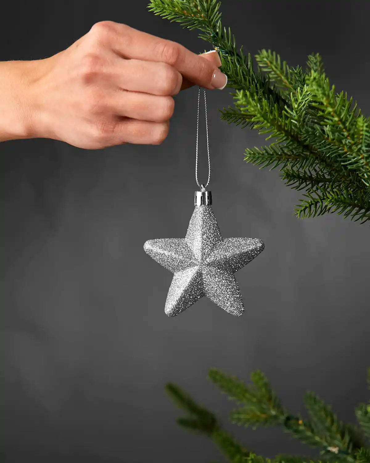Hand holding a silver 7cm glitter star decoration beside a Christmas tree.