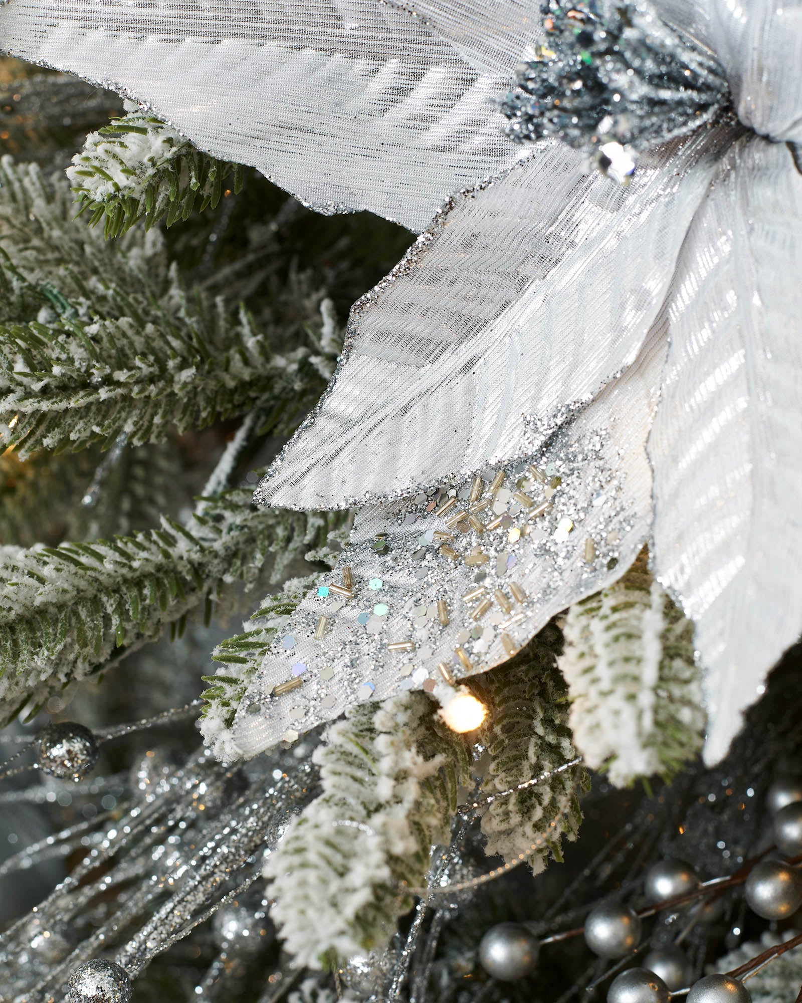 Detail view of a 30 cm silver artificial poinsettia flower with layered fabric petals, glitter edging and frosted tree branches.