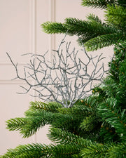 A silver glittery branch tree ornament with delicate twig-like branches, shown nestled among the green needles of a Christmas tree against a light beige paneled wall.