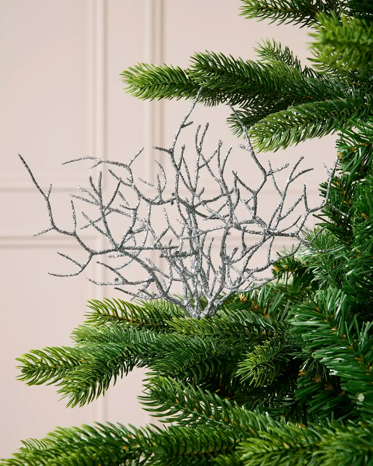 A silver glittery branch tree ornament with delicate twig-like branches, shown nestled among the green needles of a Christmas tree against a light beige paneled wall.