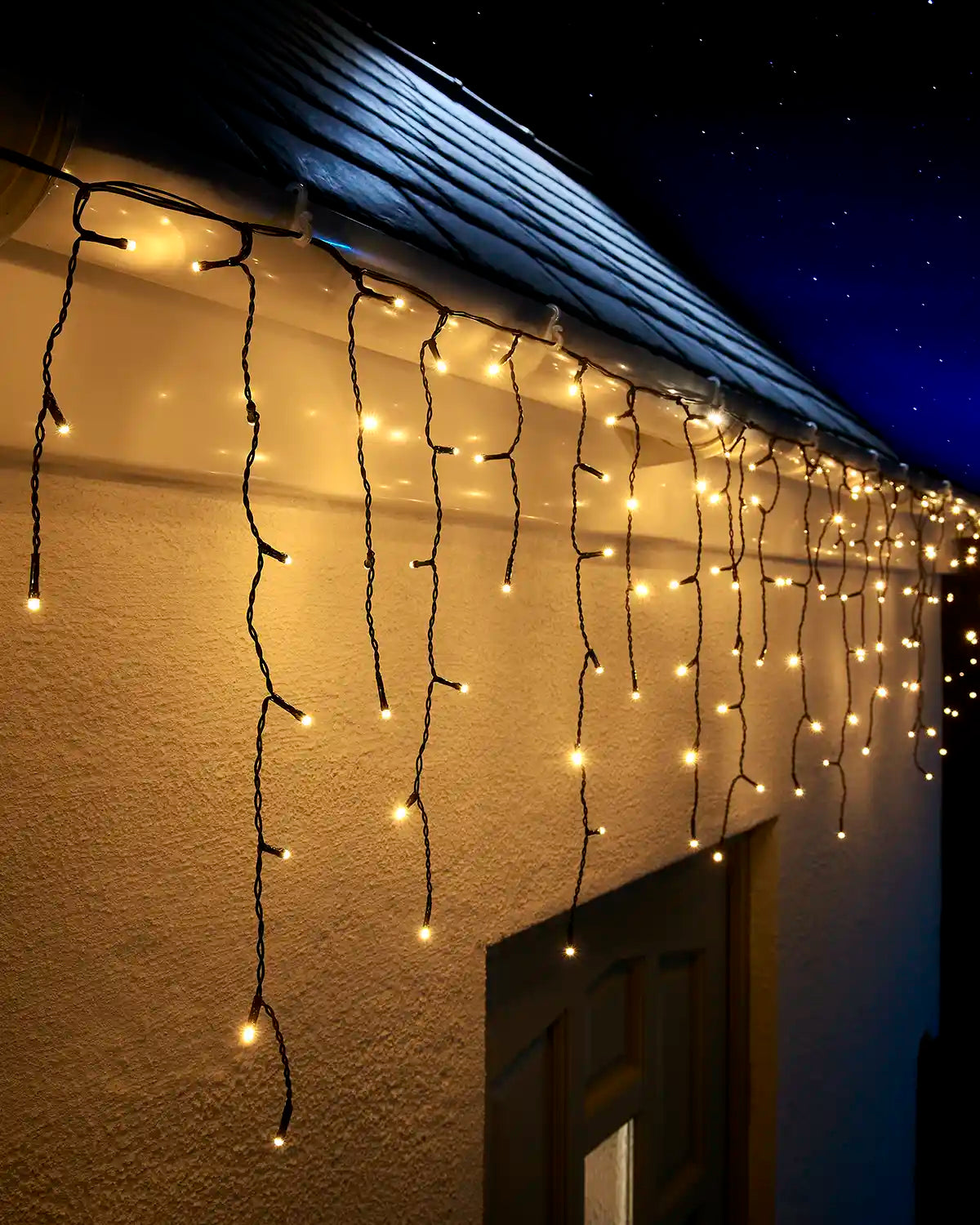 Warm white icicle lights on black cable hanging along a roofline above an exterior wall and doorway at night.