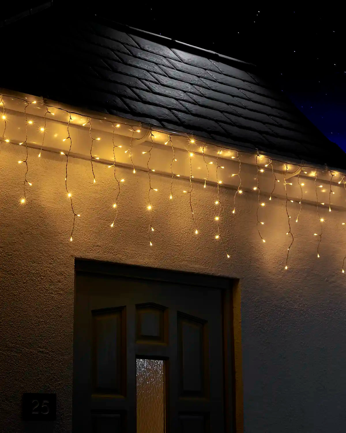 Warm white icicle lights on white cable displayed along the roofline above a front door at night.