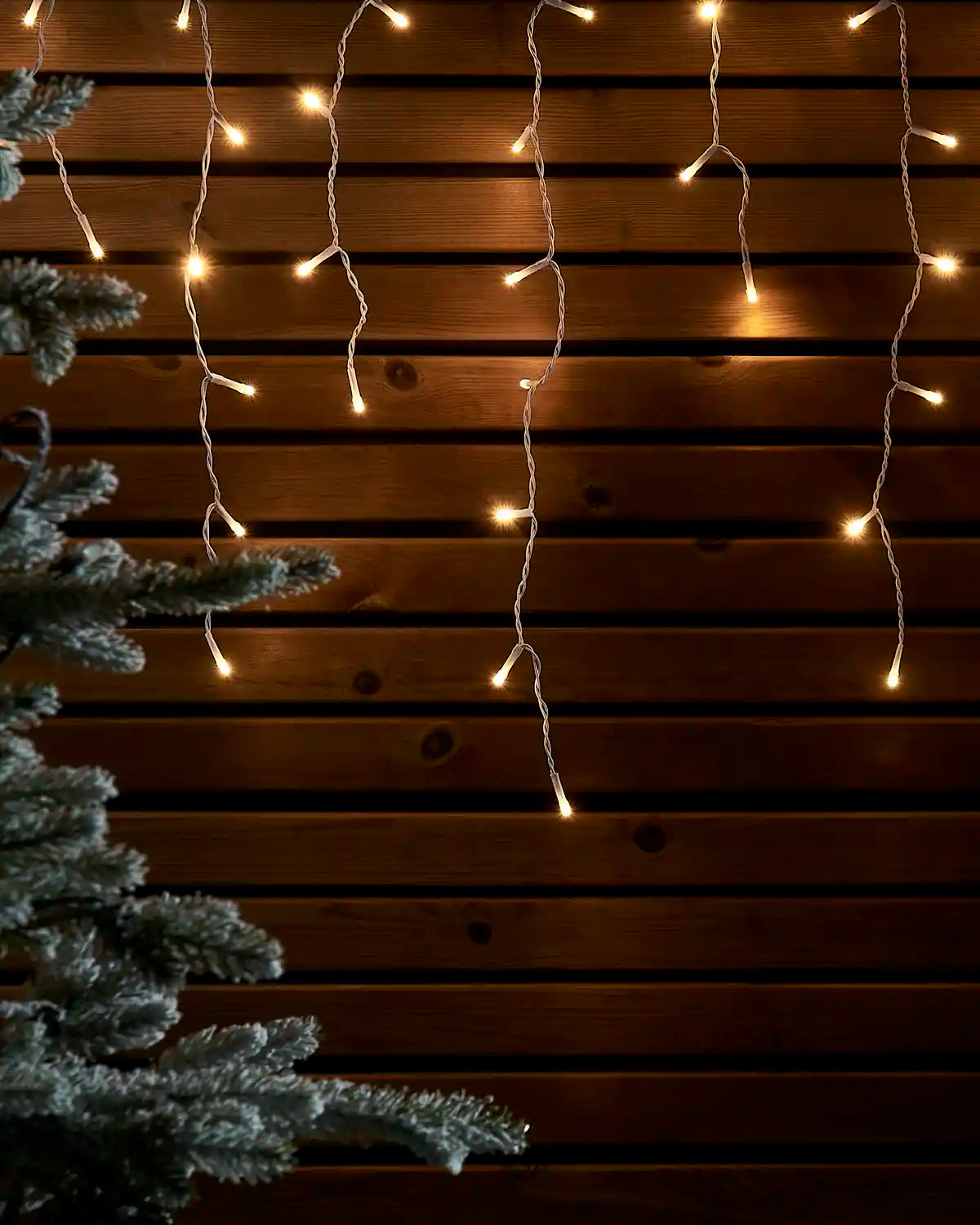 Warm white icicle lights on white cable hanging against a wooden wall beside a frosted tree.