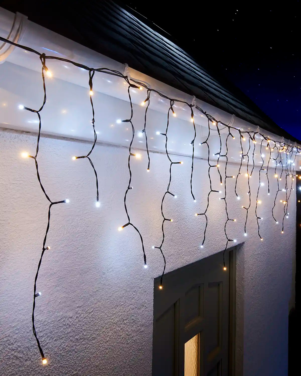 Warm white and white icicle lights on black cable displayed along an outdoor roofline at night.