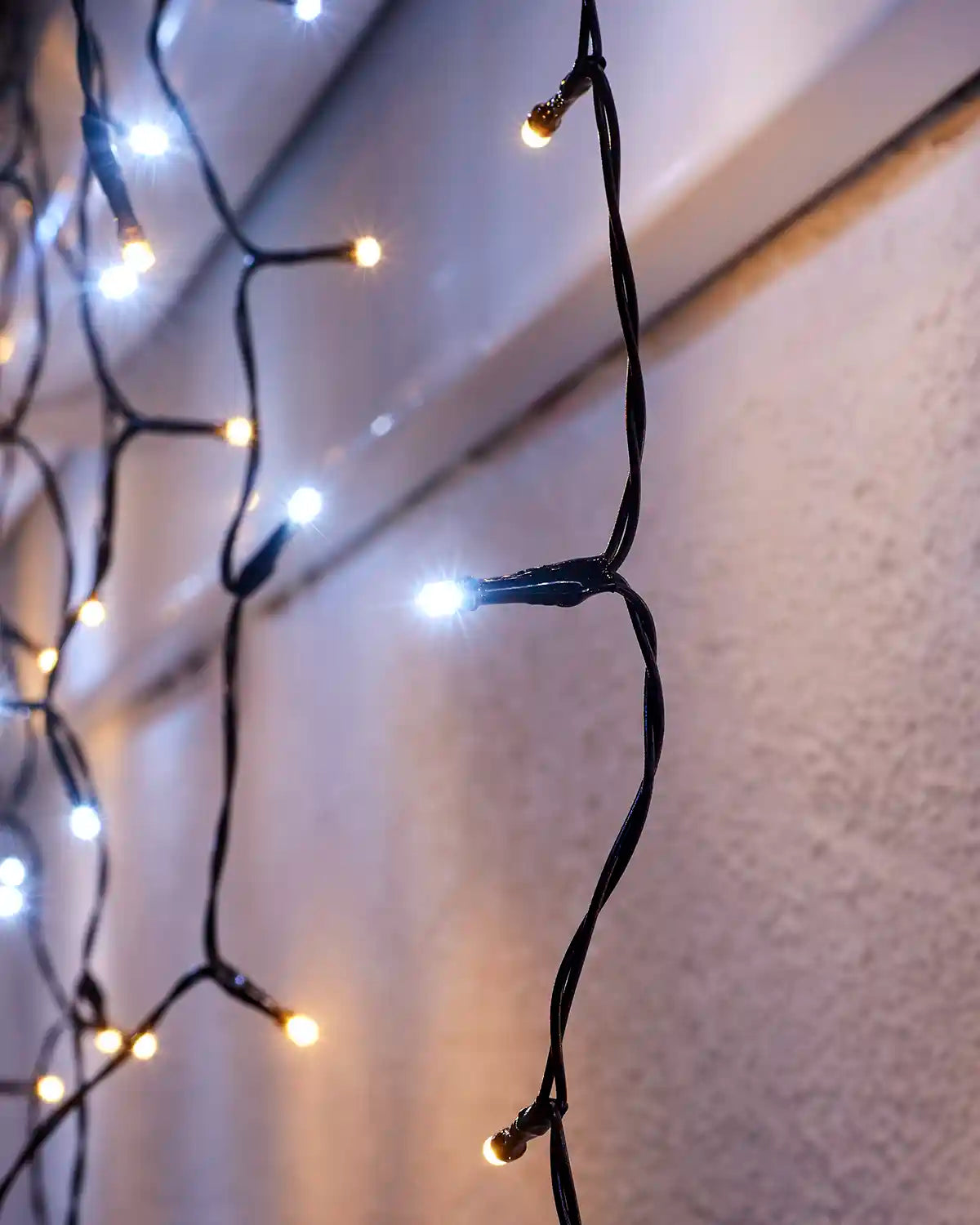 Close-up of warm white and white icicle lights on black cable against an exterior wall.