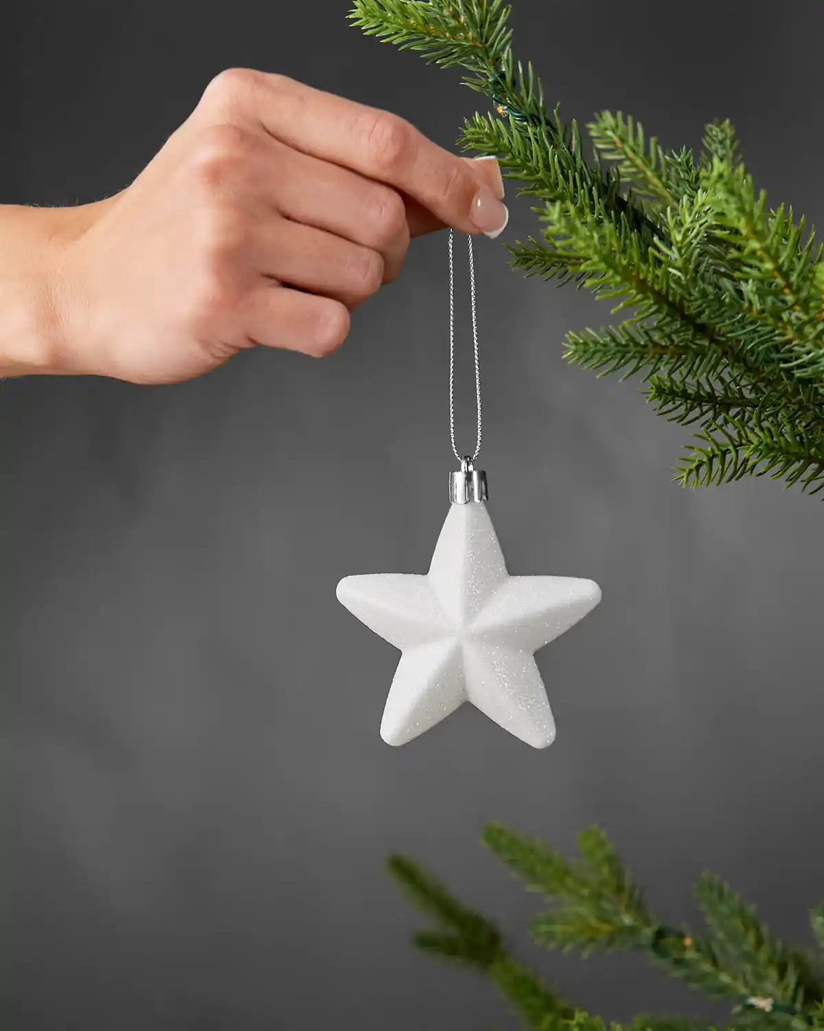 Hand holding a white 7cm glitter star ornament beside a Christmas tree.
