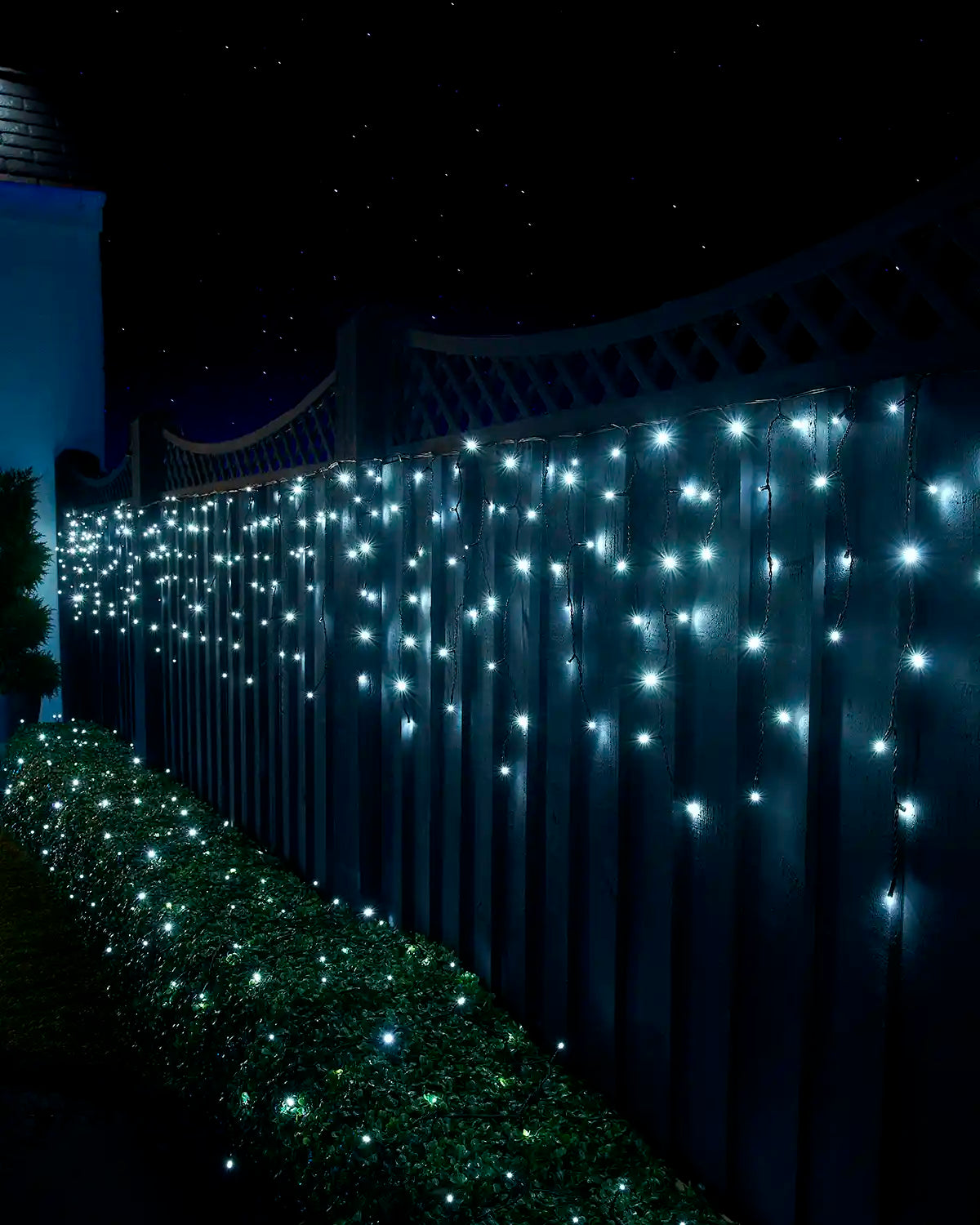 White icicle lights on black cable displayed along a garden fence above a lit hedge at night.