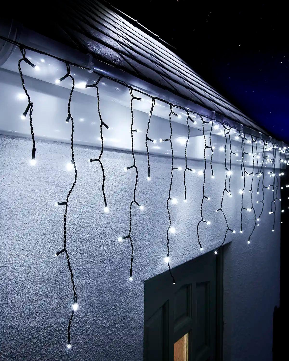 White icicle lights on black cable hanging along a roofline above an exterior wall and doorway at night.