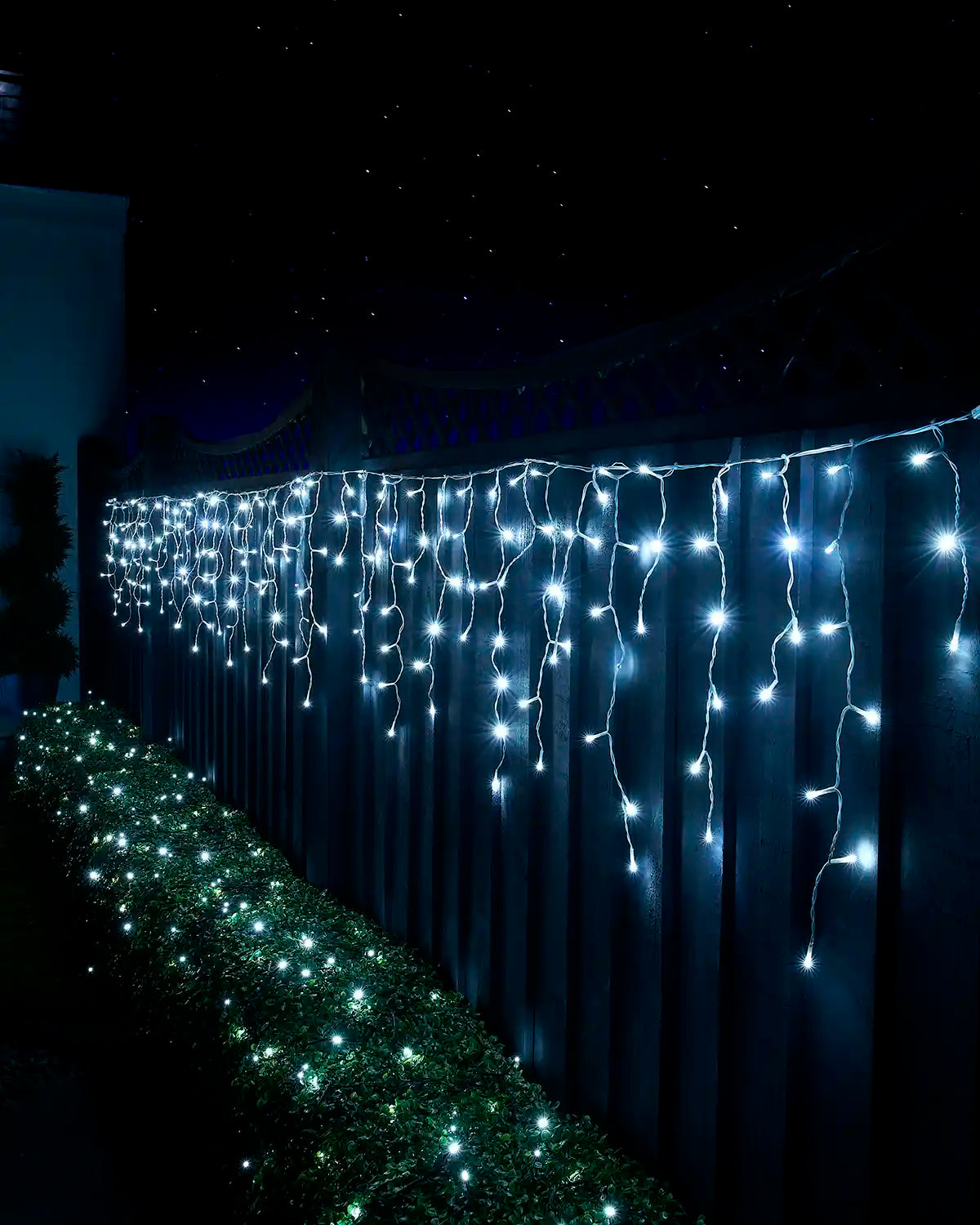 White icicle lights on white cable displayed along a garden fence above a lit hedge at night.