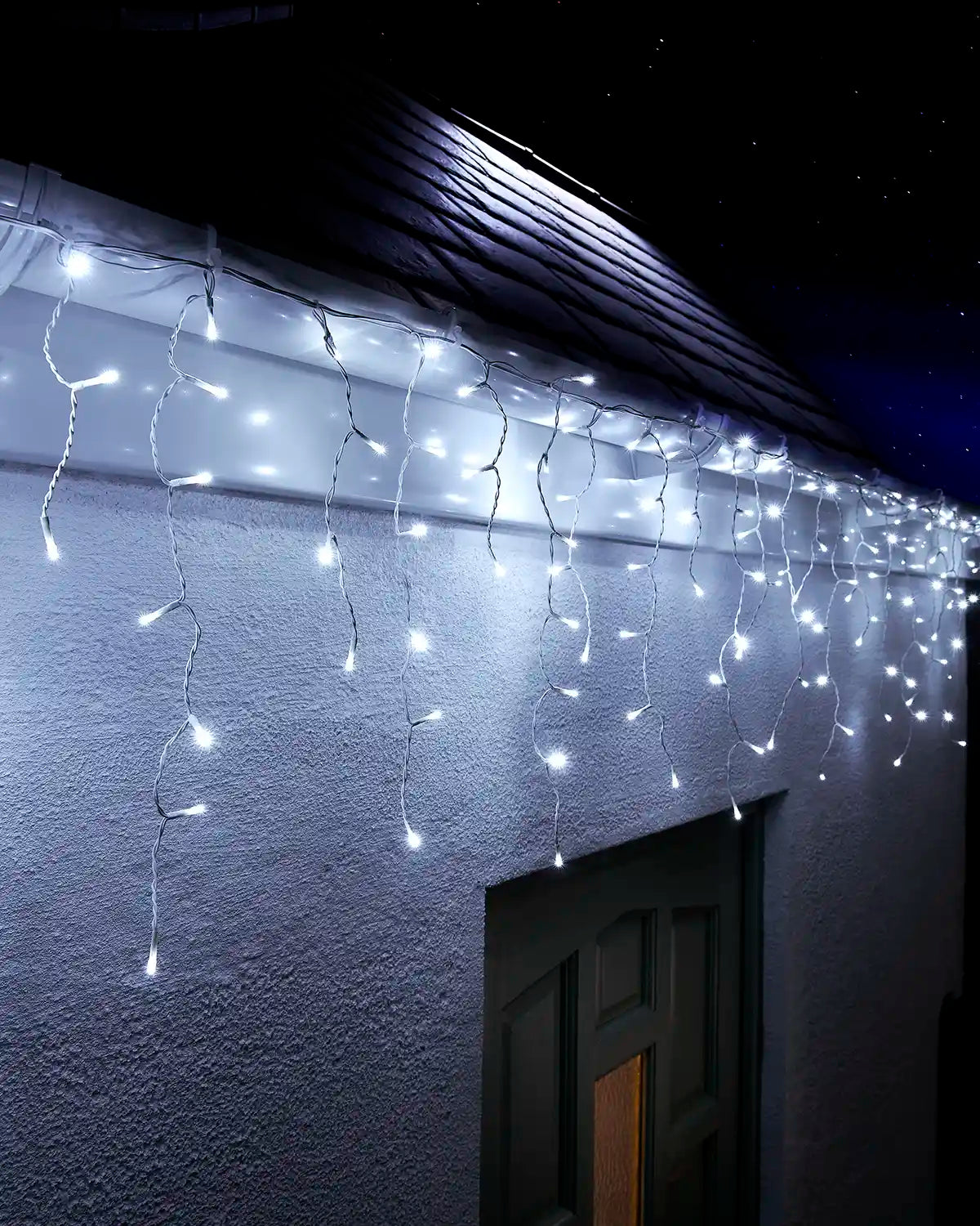 White icicle lights on white cable hanging above an exterior wall and doorway at night.