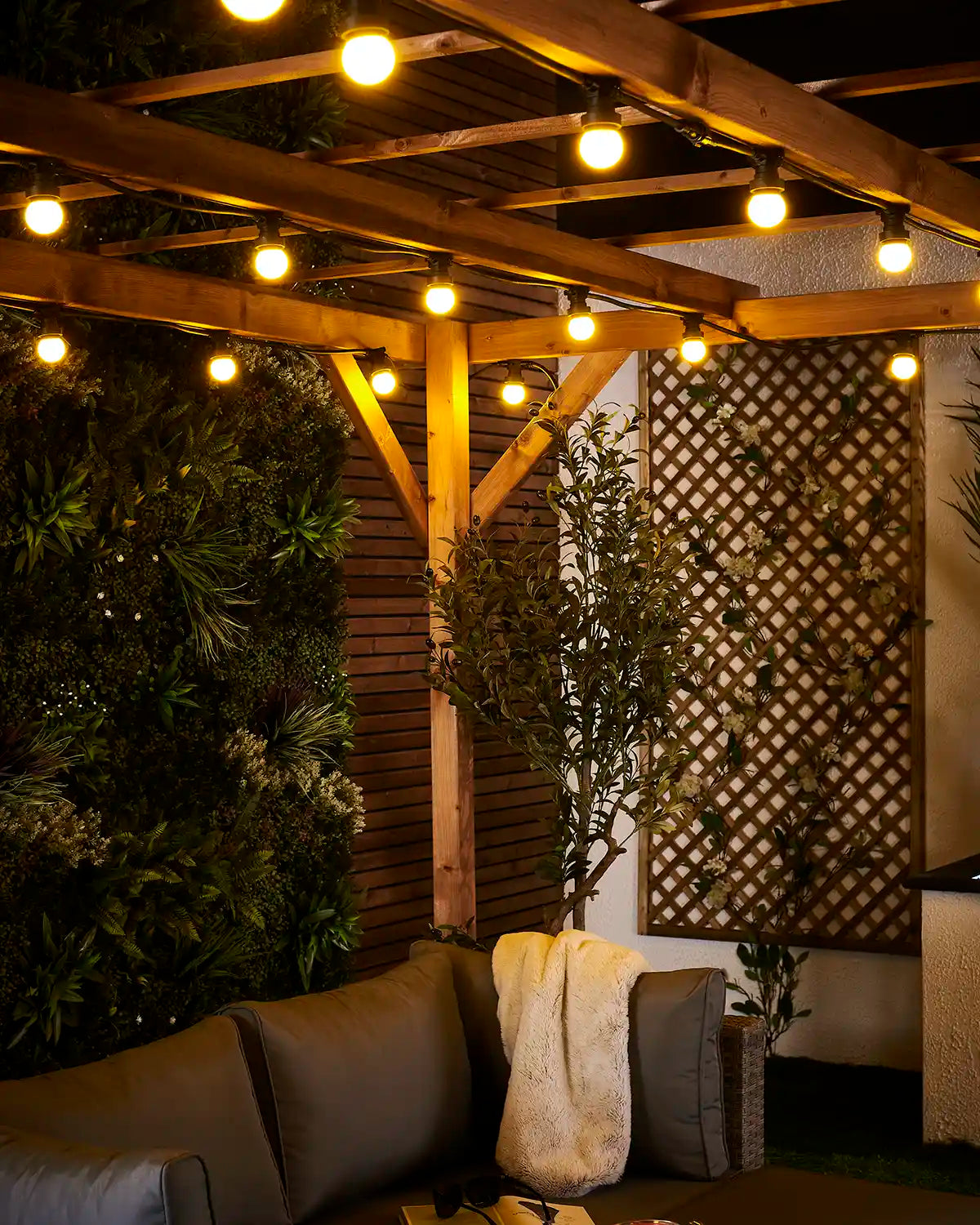 Close-up of yellow E27 festoon bulbs glowing beneath a wooden pergola with outdoor seating below.