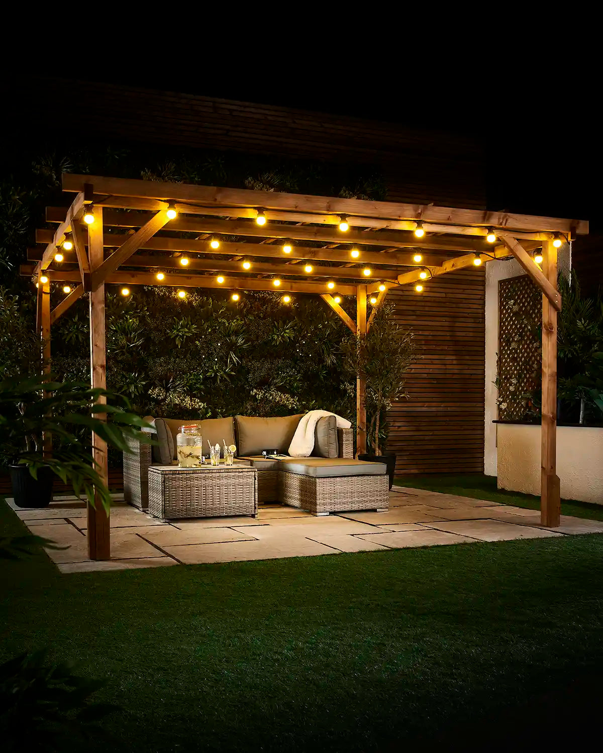 Yellow E27 festoon bulbs displayed across an outdoor wooden pergola at night with patio seating and garden plants.