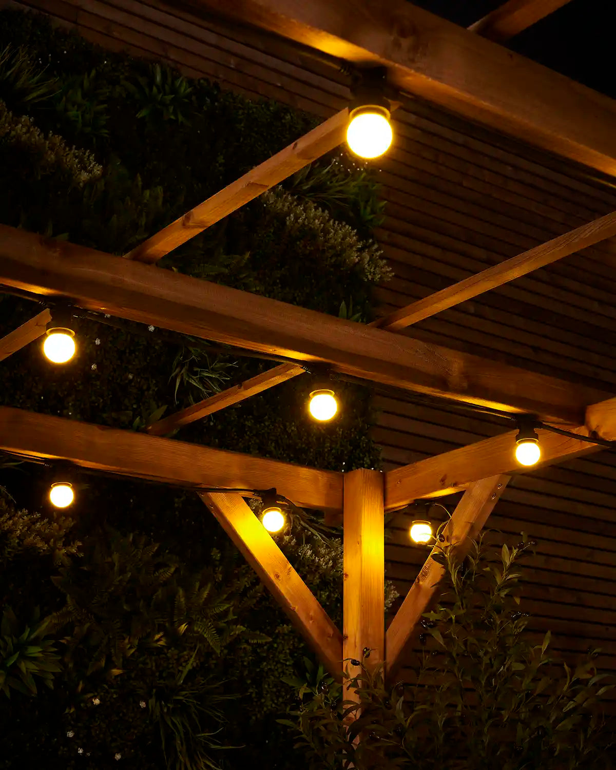 Yellow E27 festoon bulbs glowing under wooden pergola beams with garden foliage in the background.