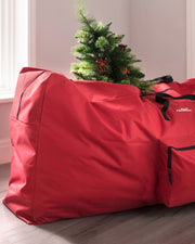 A red Christmas tree storage bag with a Christmas tree inside it, placed on the floor against a wall.