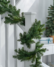 A green garland with warm white LED lights draped over a staircase, with a white wall and decorative accents in the background.