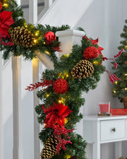 A festive pre-lit red decorated extra thick garland with warm white LED lights, pine cones, and red ornaments draped across a banister.