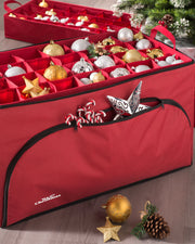 A red, fabric ornament storage box opened to display rows of Christmas ornaments inside. The box has a zipper and seems to be placed on a wooden floor with a decorated Christmas tree in the background.