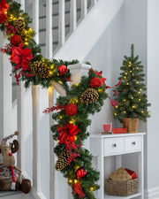 A festive pre-lit red decorated extra thick garland with warm white LED lights, pine cones, and red ornaments draped across a banister.