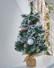 A 2-foot pre-decorated artificial Christmas tree with snow-flocked green branches, red berries, pinecones, baubles, and flowing ribbons, set in a rustic hessian-wrapped base.
