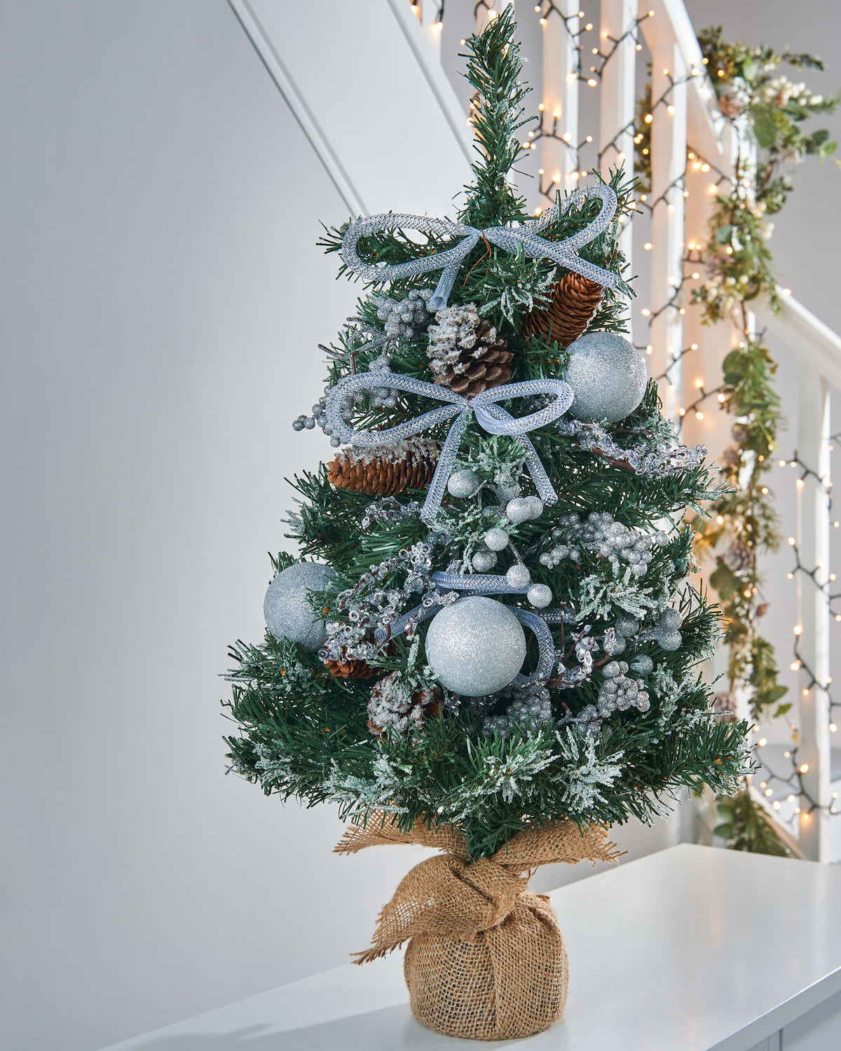 A 2-foot pre-decorated artificial Christmas tree with snow-flocked green branches, red berries, pinecones, baubles, and flowing ribbons, set in a rustic hessian-wrapped base.