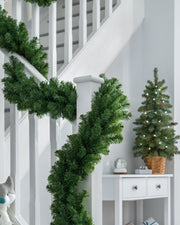 A green, extra thick pine garland draped around a staircase banister, with a white railing and a decorated Christmas tree in the background.