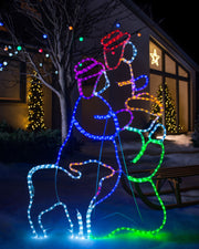 A neon rope light silhouette depicting three Wise Men and a sheep, illuminated with multi-colored LED lights, displayed outdoors at night.