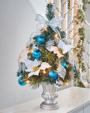 Pre-lit potted decorated Christmas tree with a silver pot, adorned with blue ornaments, white flowers, and green leaves, illuminated with warm white LED lights.