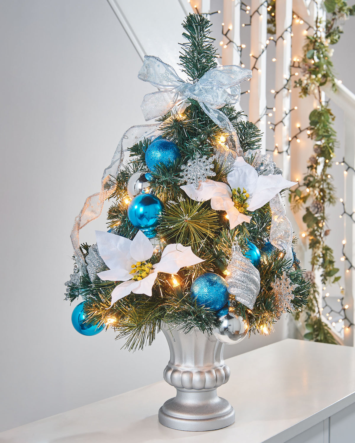 Pre-lit potted decorated Christmas tree with a silver pot, adorned with blue ornaments, white flowers, and green leaves, illuminated with warm white LED lights.