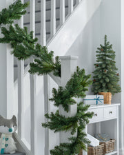 A green garland with warm white LED lights draped over a staircase, with a white wall and decorative accents in the background.