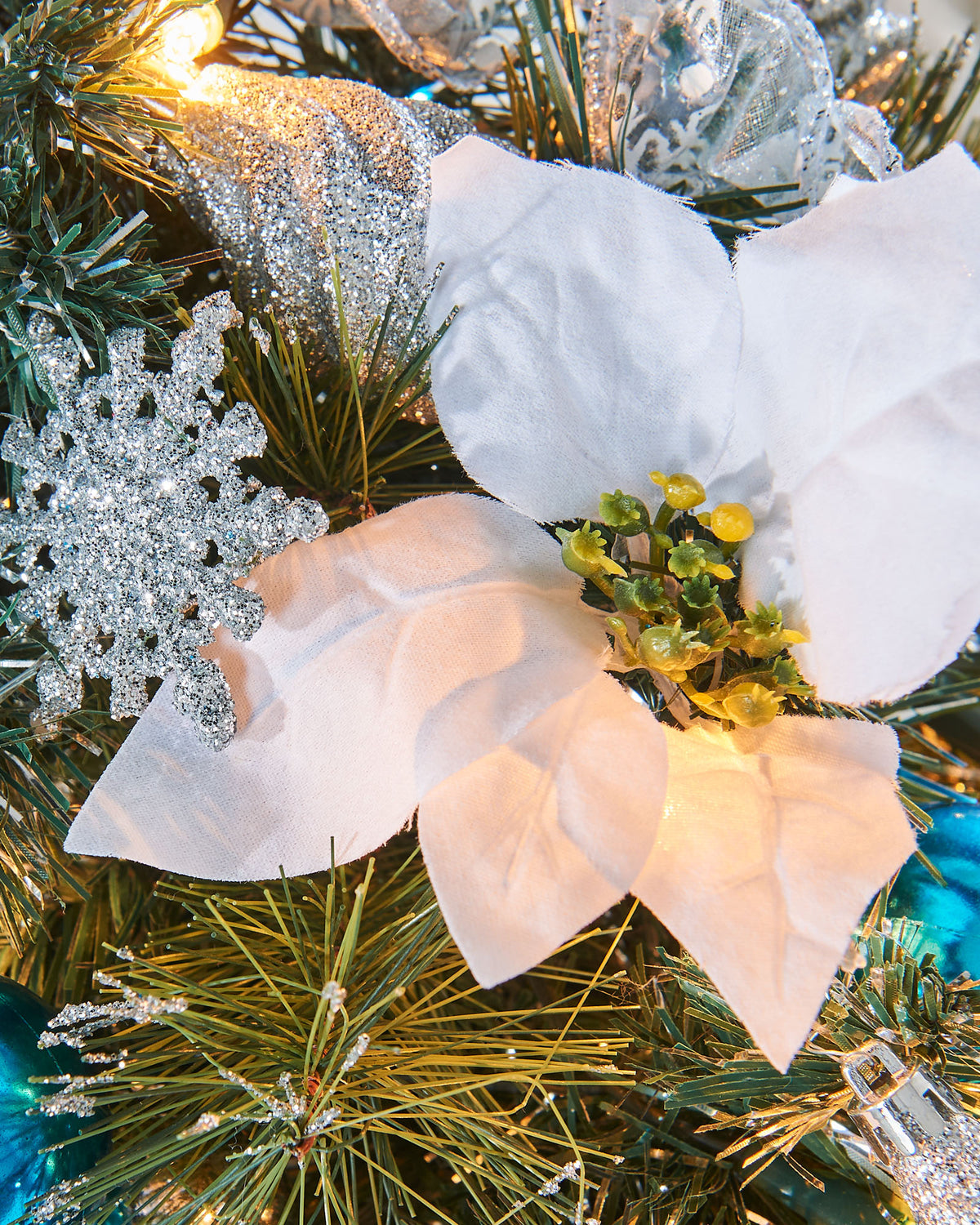 Pre-lit potted decorated Christmas tree with a silver pot, adorned with blue ornaments, white flowers, and green leaves, illuminated with warm white LED lights.
