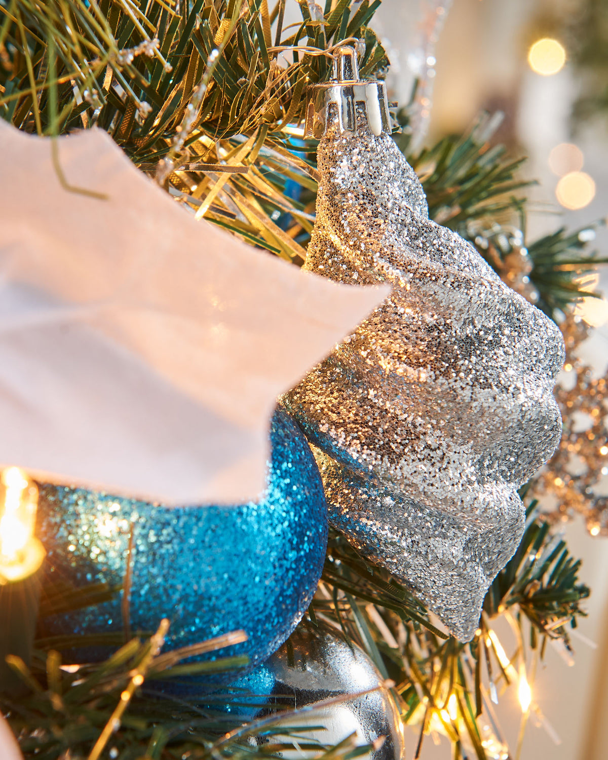 Pre-lit potted decorated Christmas tree with a silver pot, adorned with blue ornaments, white flowers, and green leaves, illuminated with warm white LED lights.