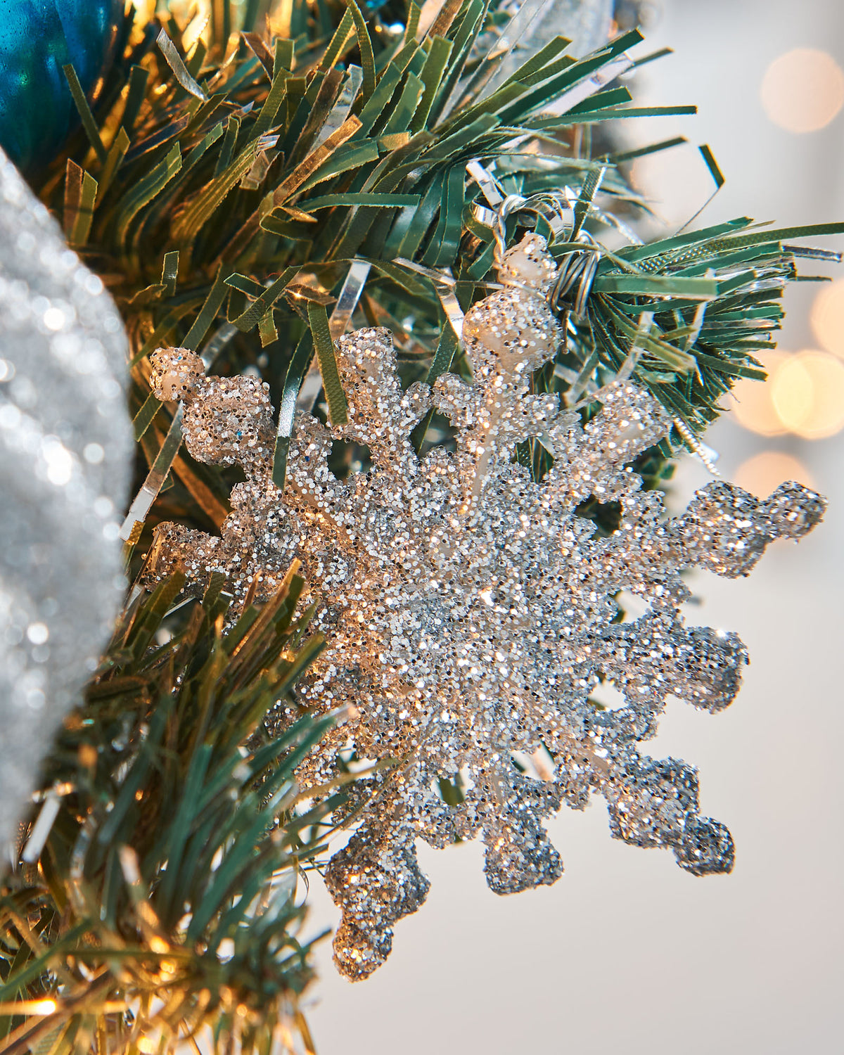 Pre-lit potted decorated Christmas tree with a silver pot, adorned with blue ornaments, white flowers, and green leaves, illuminated with warm white LED lights.