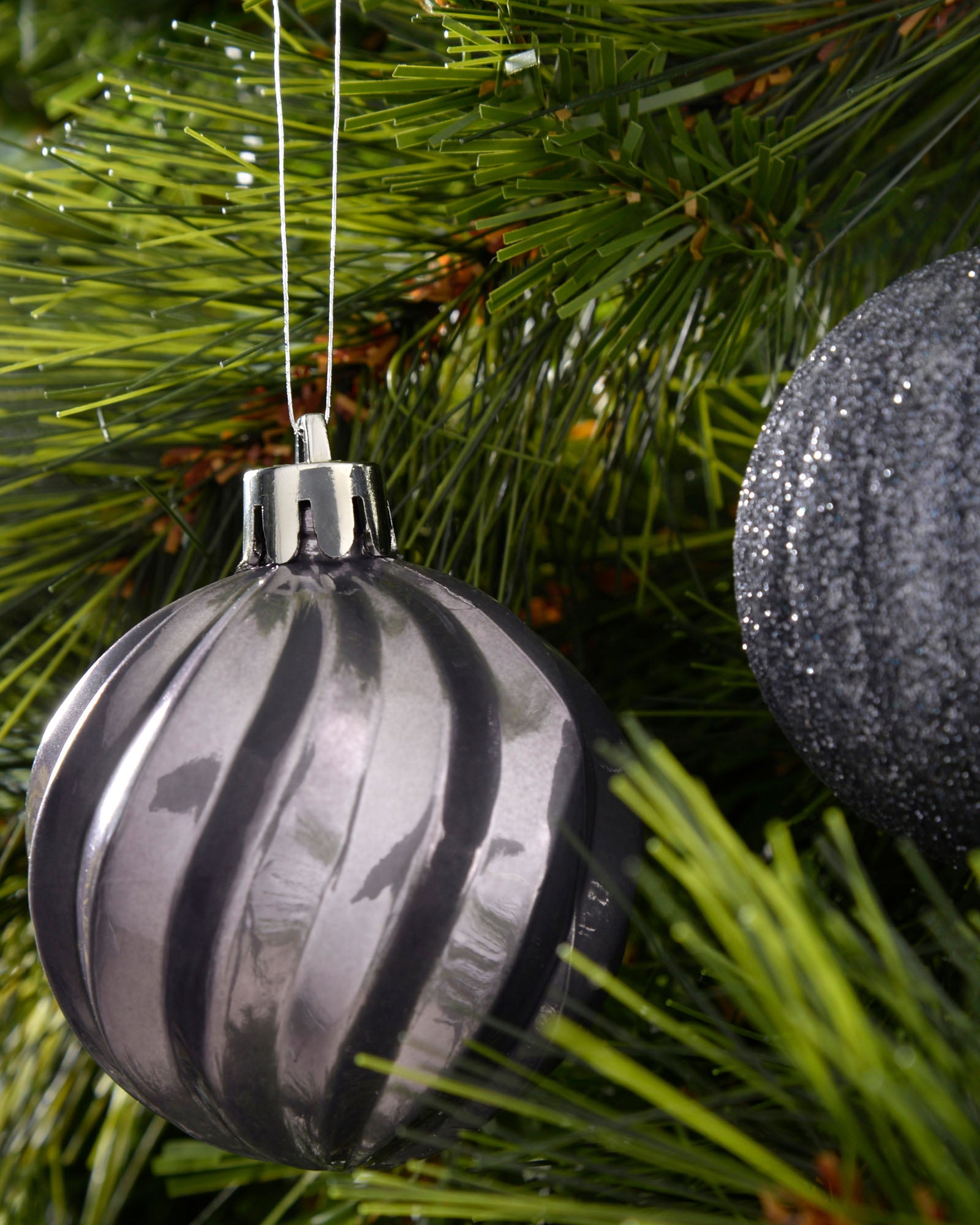 Grey ribbed 6cm shatterproof bauble hanging on a green Christmas tree beside a dark glitter bauble